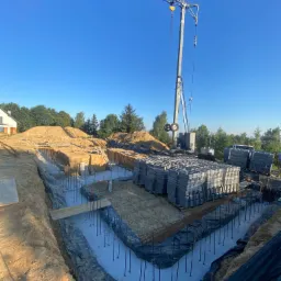 Wykonany wykop pod fundamenty z widocznym zbrojeniem i izolacją, na placu budowy stoi żuraw i palety z bloczkami betonowymi, w tle drzewa i dom.