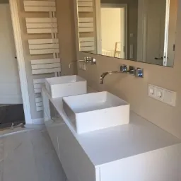 Dwie nowoczesne, kwadratowe umywalki nablatowe zamontowane na białej szafce łazienkowej z chromowanymi bateriami, widoczne gniazdka elektryczne na ścianie i drabinkowy grzejnik w tle.