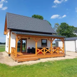 Gotowy, biały dom z drewnianą werandą i grafitowym dachem na tle błękitnego nieba.