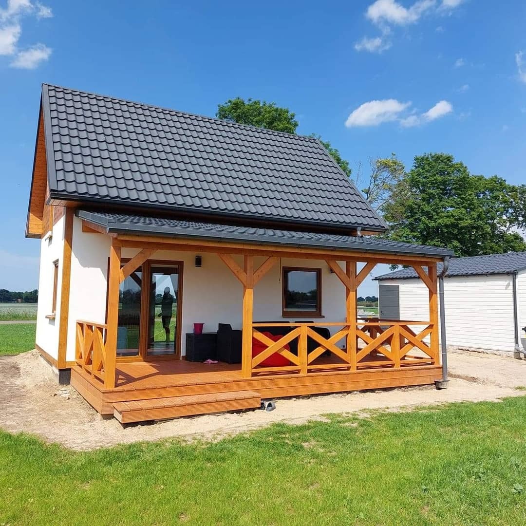 Gotowy, biały dom z drewnianą werandą i grafitowym dachem na tle błękitnego nieba.