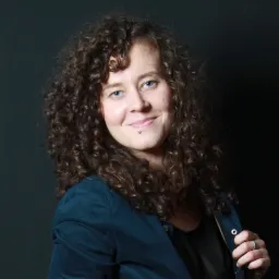 Portret uśmiechniętej kobiety z burzą kręconych włosów, ubrana w granatową marynarkę, pozująca na ciemnym tle.