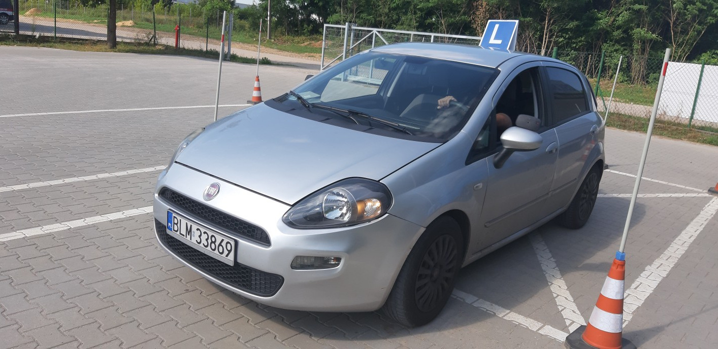 Srebrny samochód z literą 'L' na dachu, zaparkowany na placu manewrowym z pachołkami i słupkami, widoczny numer rejestracyjny BLM 33869.