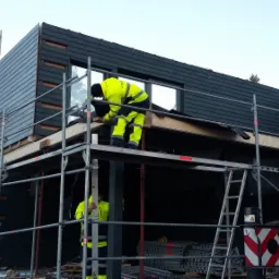 Budowa domu szkieletowego z ciemną elewacją drewnianą: dwóch pracowników w odblaskowych ubraniach montuje elementy konstrukcyjne na rusztowaniu.