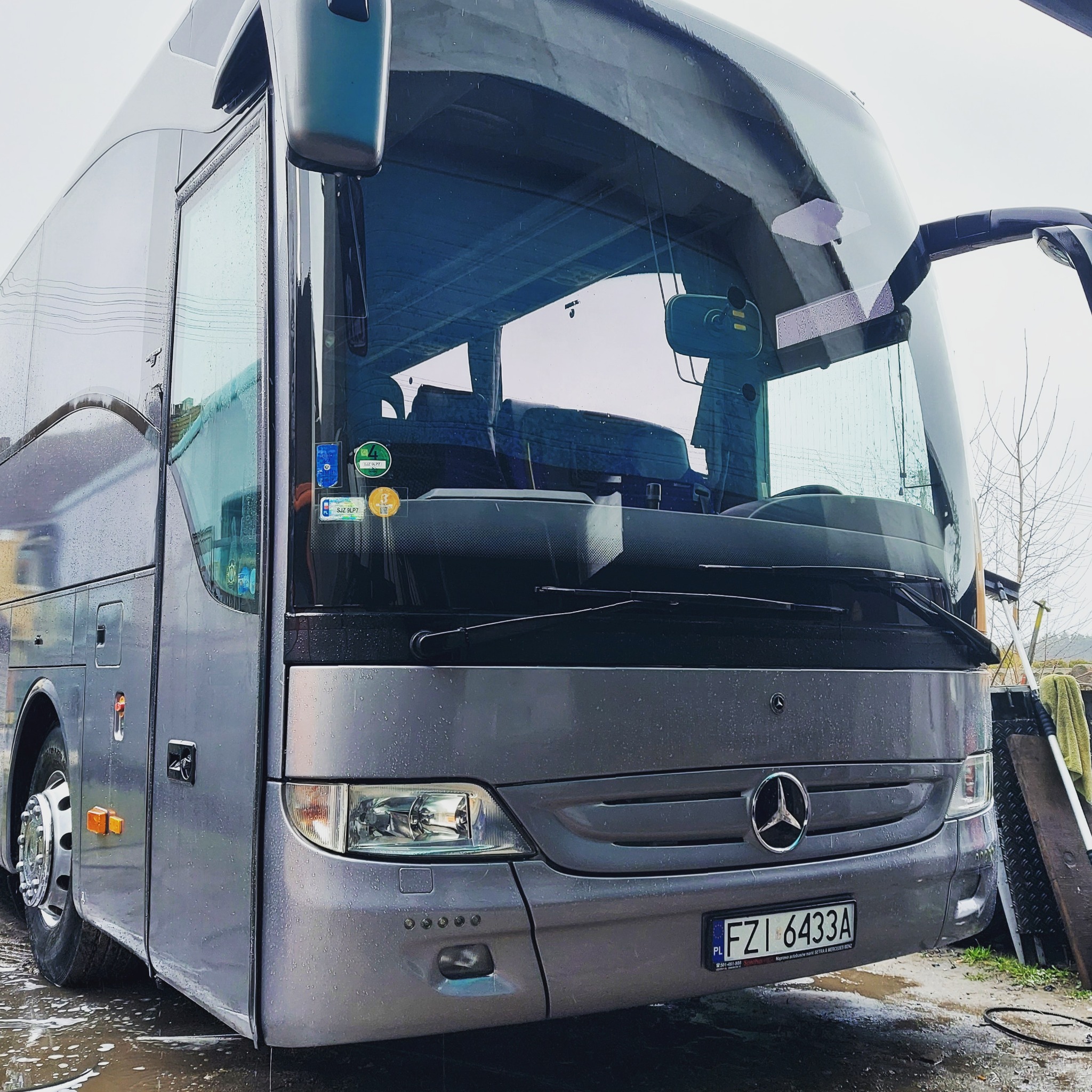 Szary autobus Mercedes-Benz z otwartymi drzwiami i mokrą karoserią, widoczne naklejki ekologiczne na szybie i tablica rejestracyjna FZI 6433A