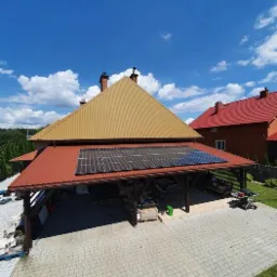 Panele słoneczne zamontowane na dachu wiaty garażowej z brązową blachodachówką, widok z góry na podjazd z kostki brukowej i sąsiedni dom z czerwoną blachodachówką w słoneczny dzień.