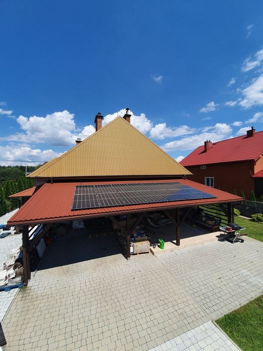 Panele słoneczne zamontowane na dachu wiaty garażowej z brązową blachodachówką, widok z góry na podjazd z kostki brukowej i sąsiedni dom z czerwoną blachodachówką w słoneczny dzień.