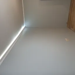 Świeżo pomalowane wnętrze z szarymi ścianami, białym wykończeniem sufitu i fragmentem parkietu na podłodze, widoczne gniazdko elektryczne na ścianie.