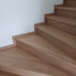 Jasne, drewniane schody z widocznym usłojeniem drewna, przylegające do białej ściany, widok z dołu.