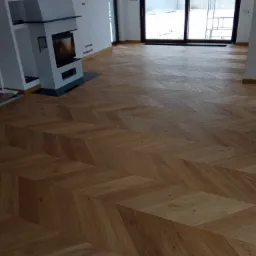 Ukończona podłoga z parkietu ułożonego we wzór jodełki w nowym, jasnym salonie z kominkiem i dużymi oknami balkonowymi.