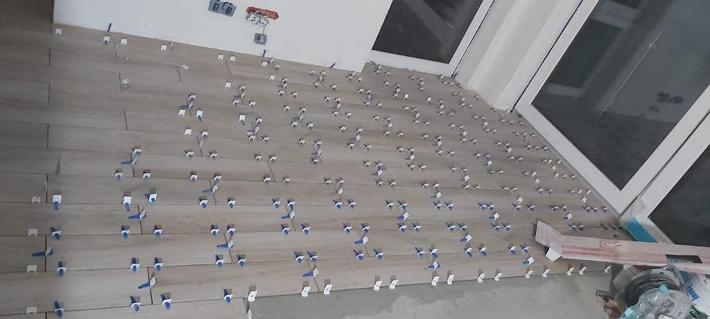 Układanie podłogi z płytek imitujących drewno, z użyciem systemu poziomowania płytek, widoczne okno balkonowe i instalacja elektryczna na ścianie.