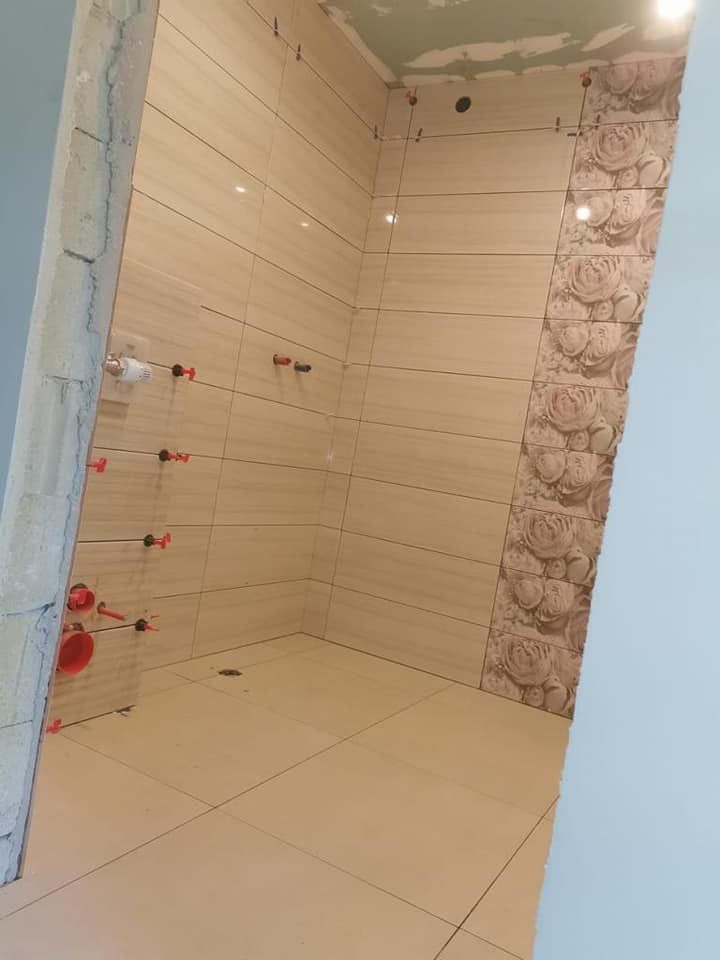 Wnętrze łazienki w trakcie układania płytek: beżowe płytki imitujące drewno na ścianach i podłodze, z dekoracyjnym pasem płytek z motywem kwiatowym, widoczne krzyżyki dystansowe i niezakończone...