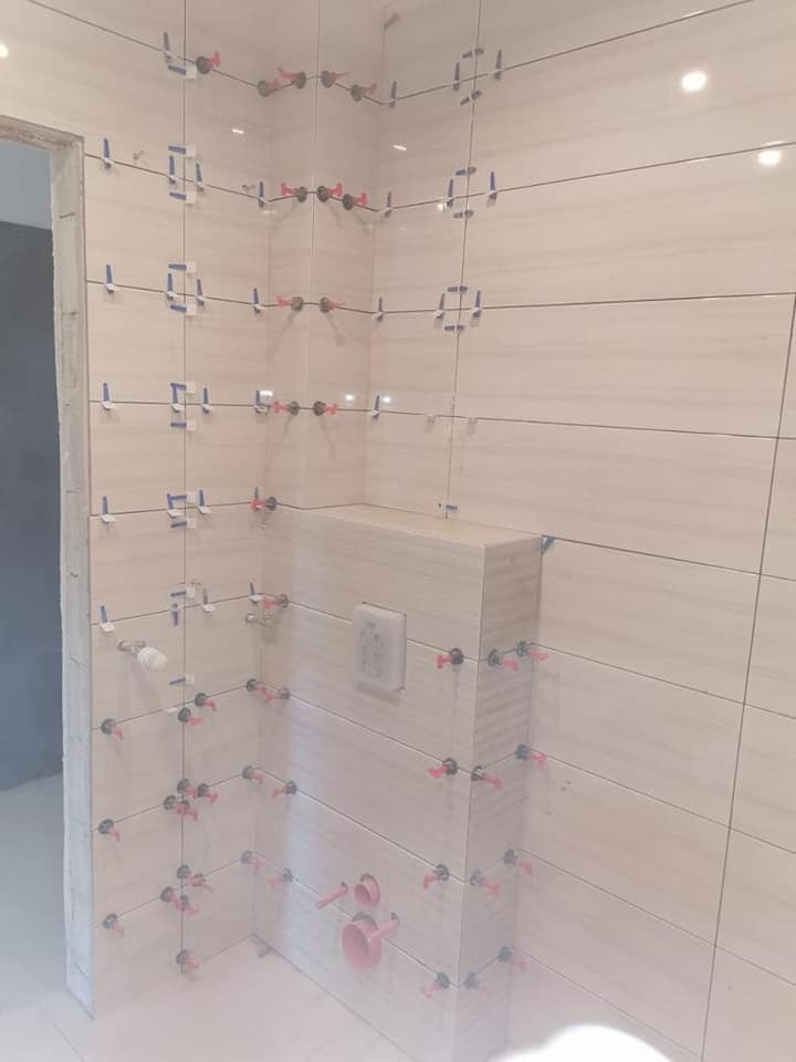 Świeżo ułożone, beżowe płytki ścienne w łazience, z widocznymi systemami poziomowania (klipsy i kliny) oraz taśmą malarską, przygotowane do fugowania; widoczne podejścia wodno-kanalizacyjne...