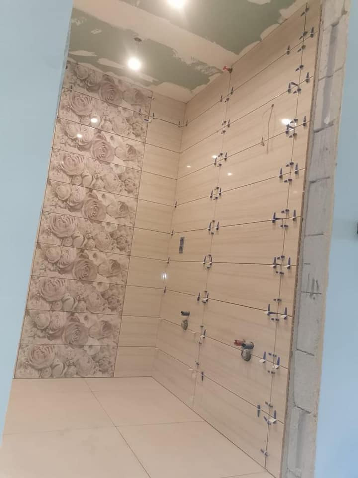 W trakcie układania beżowych płytek ściennych z motywem kwiatowym w łazience, widoczne krzyżyki dystansowe i fragment surowego sufitu.