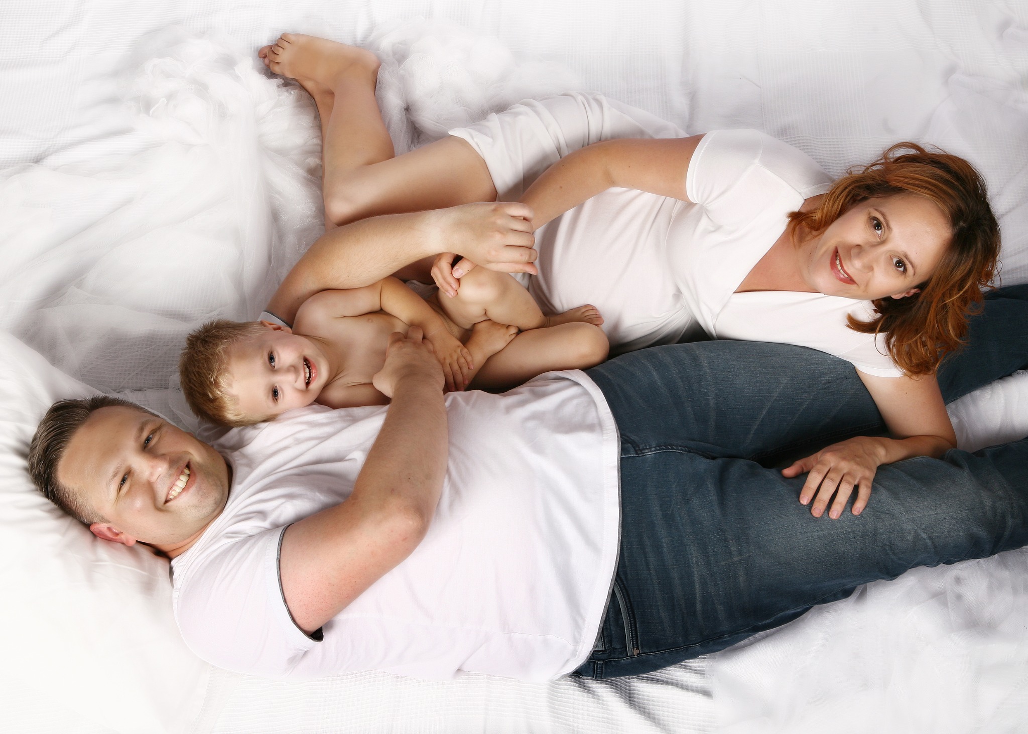 Rodzina leżąca na białej pościeli: uśmiechnięty tata, mama i syn, wszyscy ubrani w białe koszulki, tata w jeansach, mama w białych spodenkach