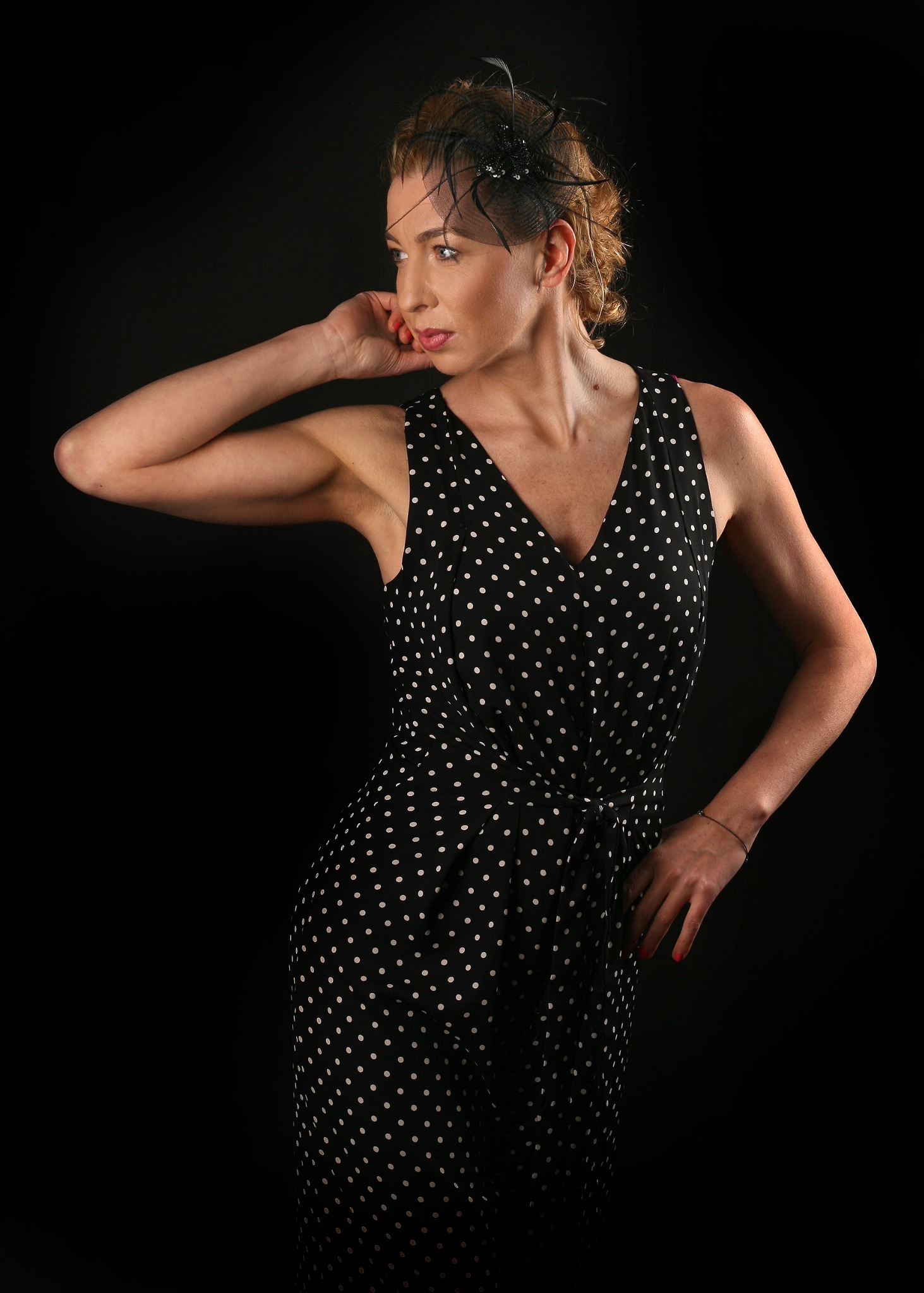 Portret kobiety w czarnej sukience w białe kropki i ozdobie we włosach, na czarnym tle, pozująca z ręką przy twarzy.
