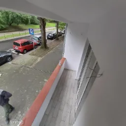 Widok z balkonu na ulicę z zaparkowanymi samochodami i chodnikiem, na balkonie jasnoszare płytki podłogowe i pomarańczowa barierka, widoczna butelka wody.