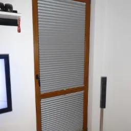 Szare plisy okienne w drewnianej ramie, zamontowane w oknie, widoczne z bliska w jasnym wnętrzu, z czarną klamką i fragmentem głośnika na srebrnej nóżce.