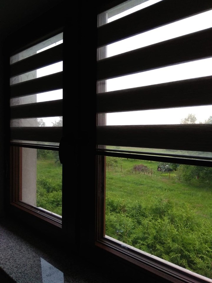 Okna z założonymi roletami zebra w ciemnym odcieniu, przez które widać zielony ogród z drzewami i traktorem w oddali.