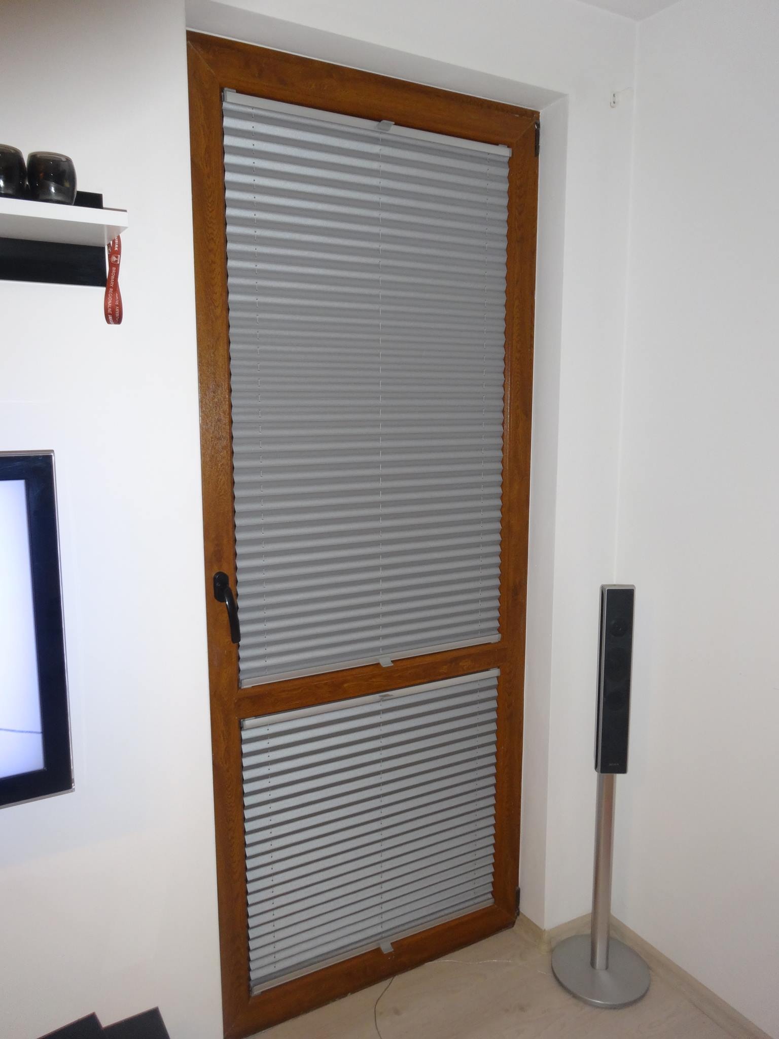 Szare plisy okienne w drewnianej ramie, zamontowane w oknie, widoczne z bliska w jasnym wnętrzu, z czarną klamką i fragmentem głośnika na srebrnej nóżce.