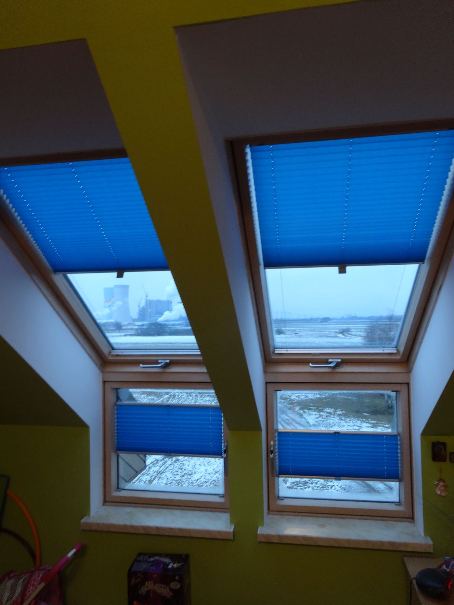 Dwa okna dachowe z zamontowanymi niebieskimi, plisowanymi osłonami, widok na zimowy krajobraz z kominami fabrycznymi w tle.