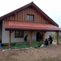 Dom w trakcie budowy z drewnianą elewacją i częściowo pokrytym czerwonym dachem, pracownicy na placu budowy, widoczny stos zielonych desek.