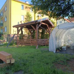 Brązowa altana z drewnianą konstrukcją w ogrodzie, obok tunel foliowy. W tle blok mieszkalny i drzewa. Na dachu altany widoczna osoba w czapce.