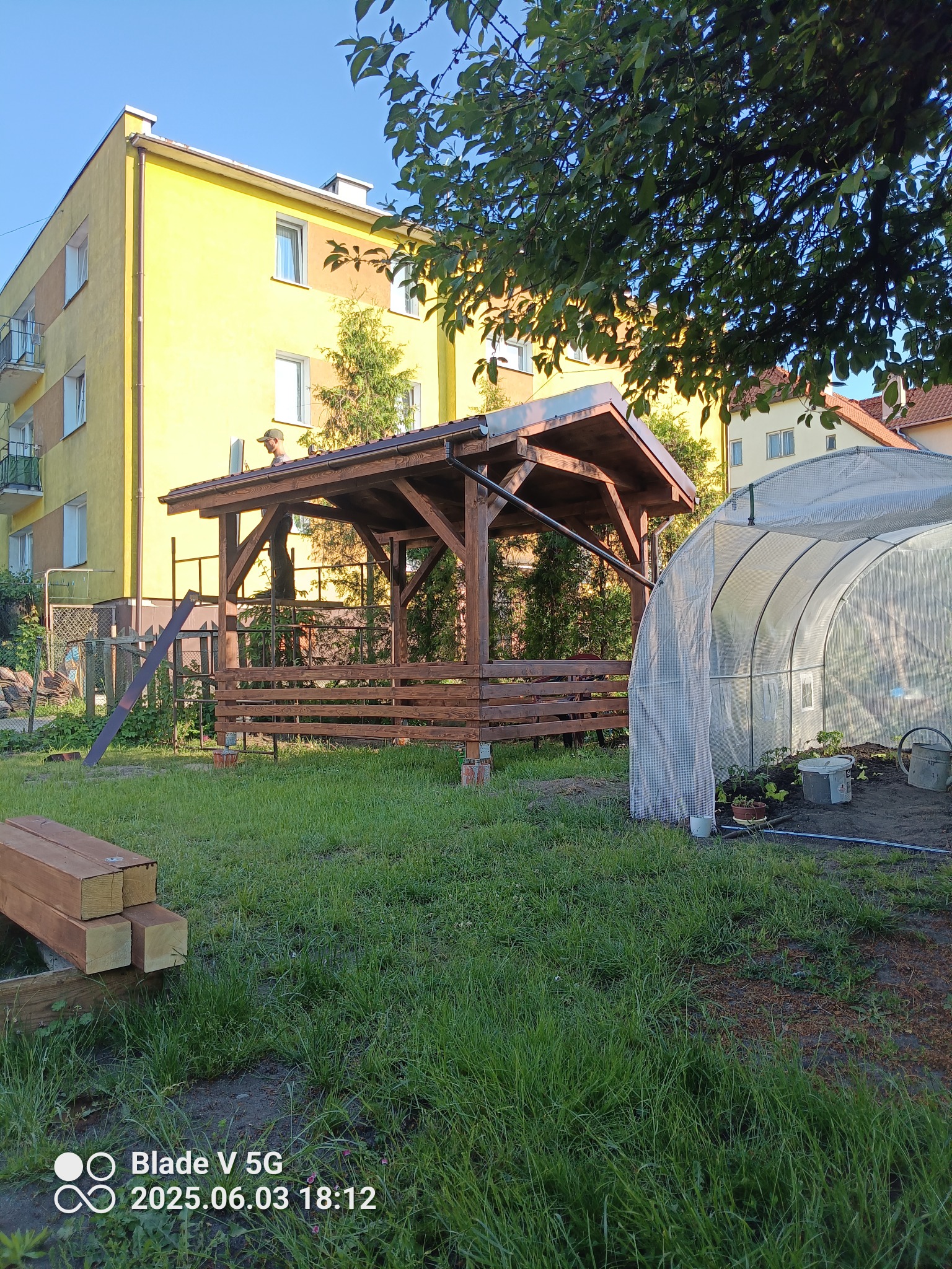 Brązowa altana z drewnianą konstrukcją w ogrodzie, obok tunel foliowy. W tle blok mieszkalny i drzewa. Na dachu altany widoczna osoba w czapce.