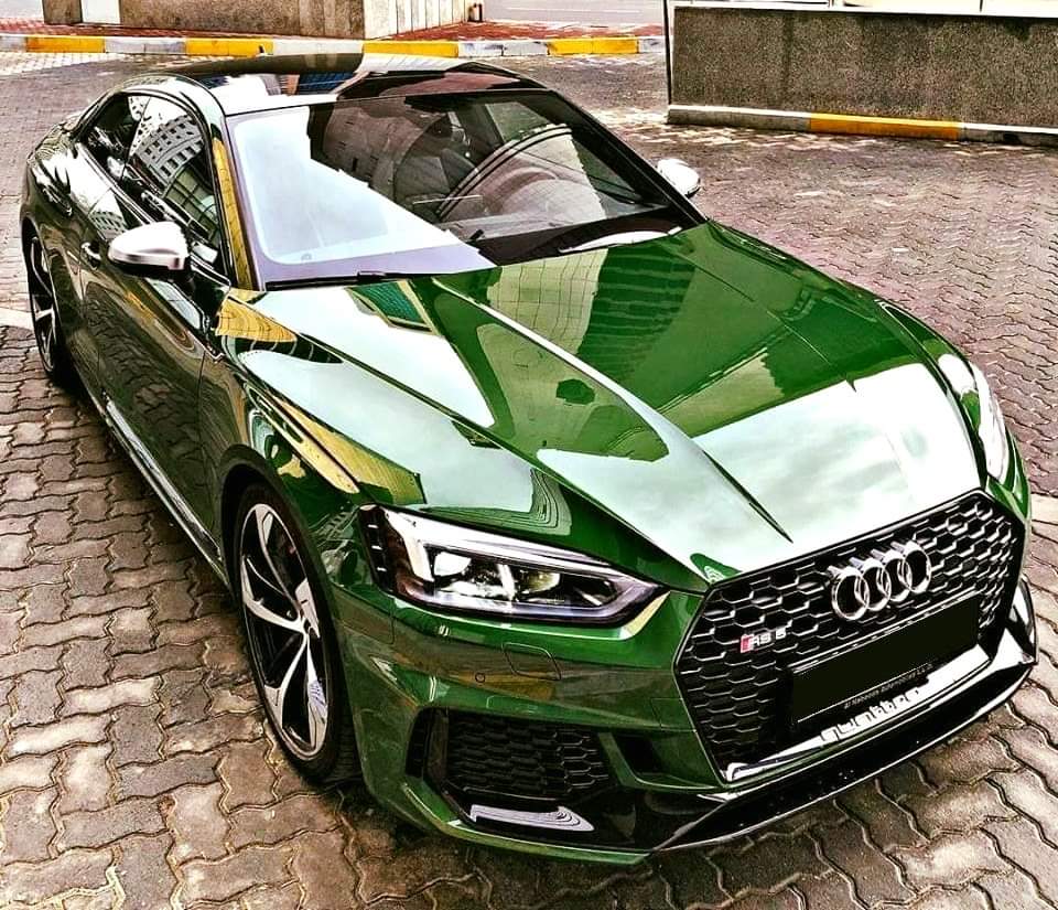 Błyszczący, zielony Audi RS5 zaparkowany na brukowanym chodniku, z odbiciem budynku w karoserii.