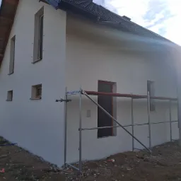 Dwukondygnacyjny dom jednorodzinny z jasną elewacją w trakcie prac wykończeniowych, widoczne rusztowania przy ścianie bocznej i zabezpieczone okna, teren wokół domu jeszcze niezagospodarowany.