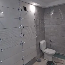 Świeżo wyłożona szara glazura z falistym wzorem w łazience, krzyżyki dystansowe w spoinach, częściowo widoczna toaleta i narzędzia na podłodze.