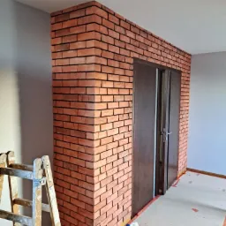 Ściana wykończona czerwoną cegłą klinkierową, obudowująca ciemne, dwuskrzydłowe drzwi w trakcie prac wykończeniowych. Na podłodze widoczne taśmy malarskie i butelka płynu do czyszczenia. Z boku...