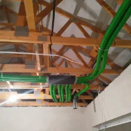 Montaż rekuperacji: centrala wentylacyjna z podłączonymi zielonymi przewodami wentylacyjnymi w surowym wnętrzu poddasza z widoczną drewnianą konstrukcją dachu.