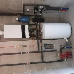 Instalacja grzewcza w kotłowni: piec gazowy Buderus, zasobnik ciepłej wody, naczynie przeponowe, rozdzielacz ogrzewania podłogowego, rury miedziane i izolowane, zamontowane na kafelkowanej ścianie.