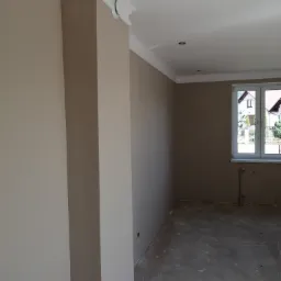 Wnętrze pokoju w trakcie remontu, widoczne świeżo pomalowane ściany w odcieniach beżu, białe listwy przysufitowe oraz surowa podłoga przygotowana do dalszych prac wykończeniowych. Przez okno...