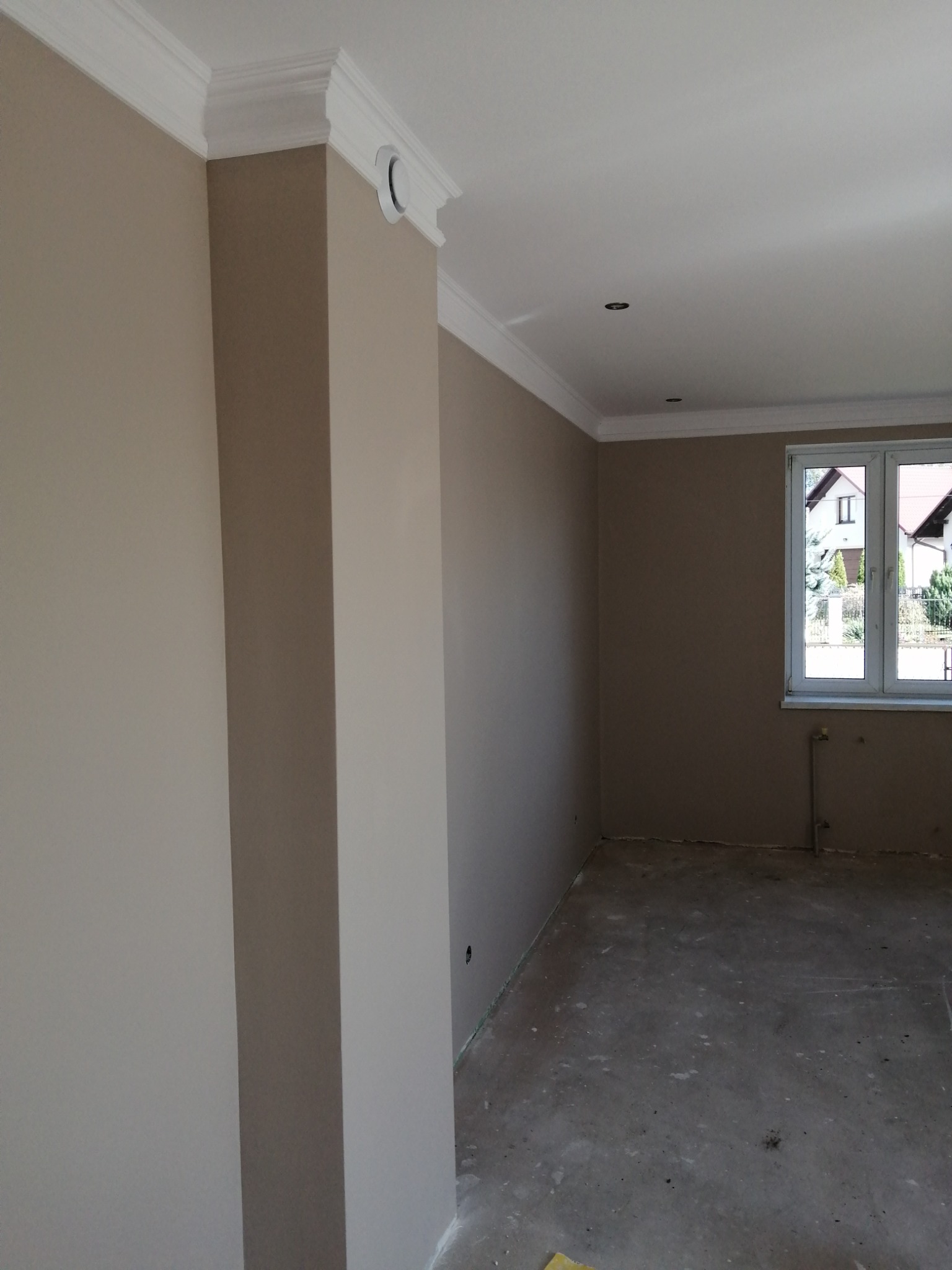 Wnętrze pokoju w trakcie remontu, widoczne świeżo pomalowane ściany w odcieniach beżu, białe listwy przysufitowe oraz surowa podłoga przygotowana do dalszych prac wykończeniowych. Przez okno...