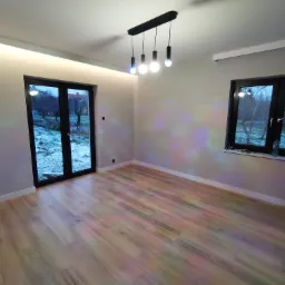 Pusty pok&oacute;j po remoncie w Wasilkowie z drewnopodobną podłogą, czarnymi ramami okiennymi i nowoczesną lampą wiszącą z czterema żar&oacute;wkami.