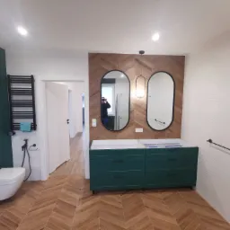 Nowoczesna łazienka w trakcie wykańczania: zielona szafka pod umywalkę bez blatu, lustra owalne z czarną ramą na ścianie z drewnopodobnych płytek, biała toaleta wisząca i grzejnik drabinkowy...