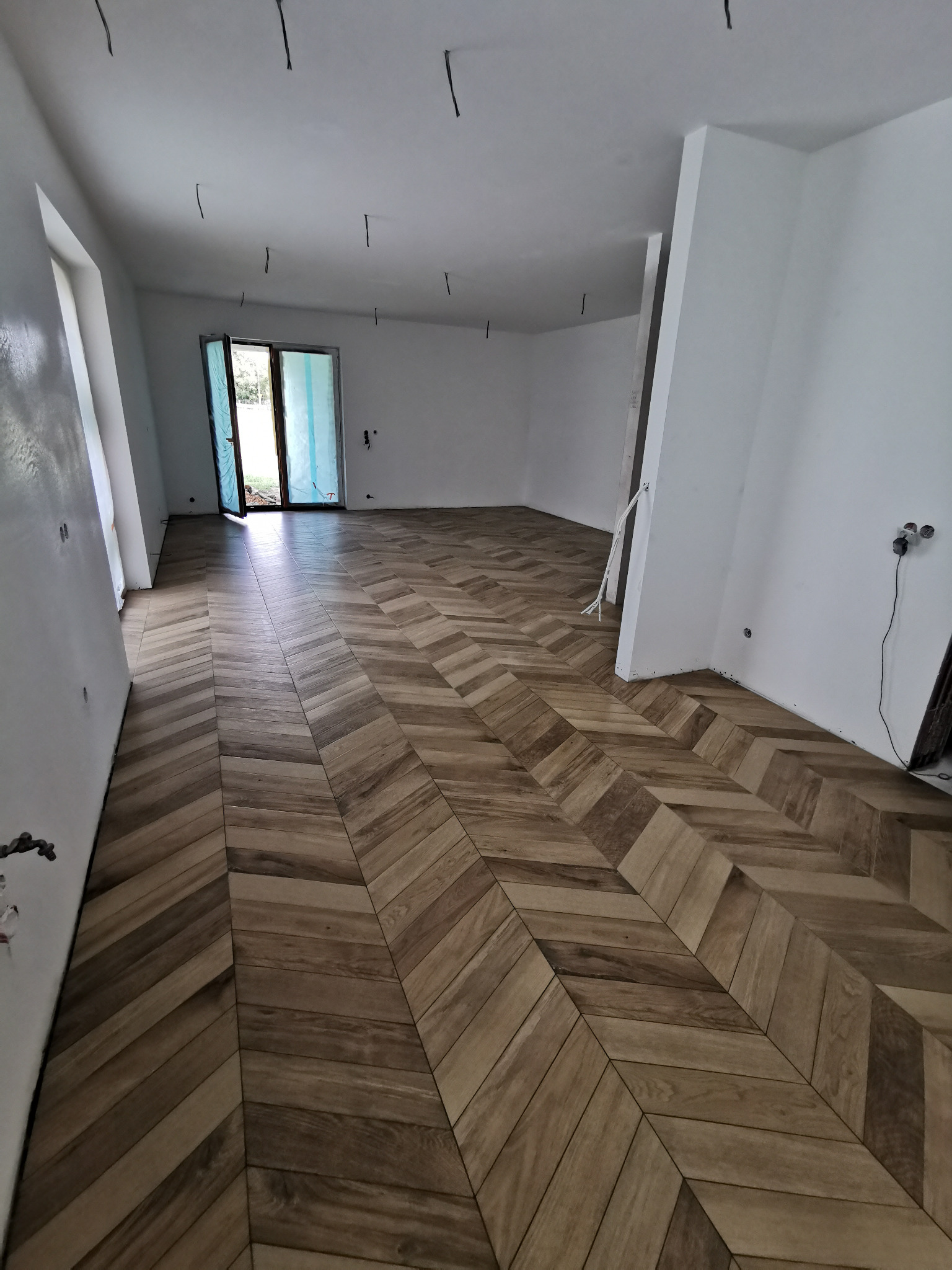 Ułożona w jodełkę, drewnopodobna podłoga w jasnym, pustym pomieszczeniu w Wasilkowie. Widoczne wystające kable ze stropu i częściowo otwarte drzwi balkonowe.