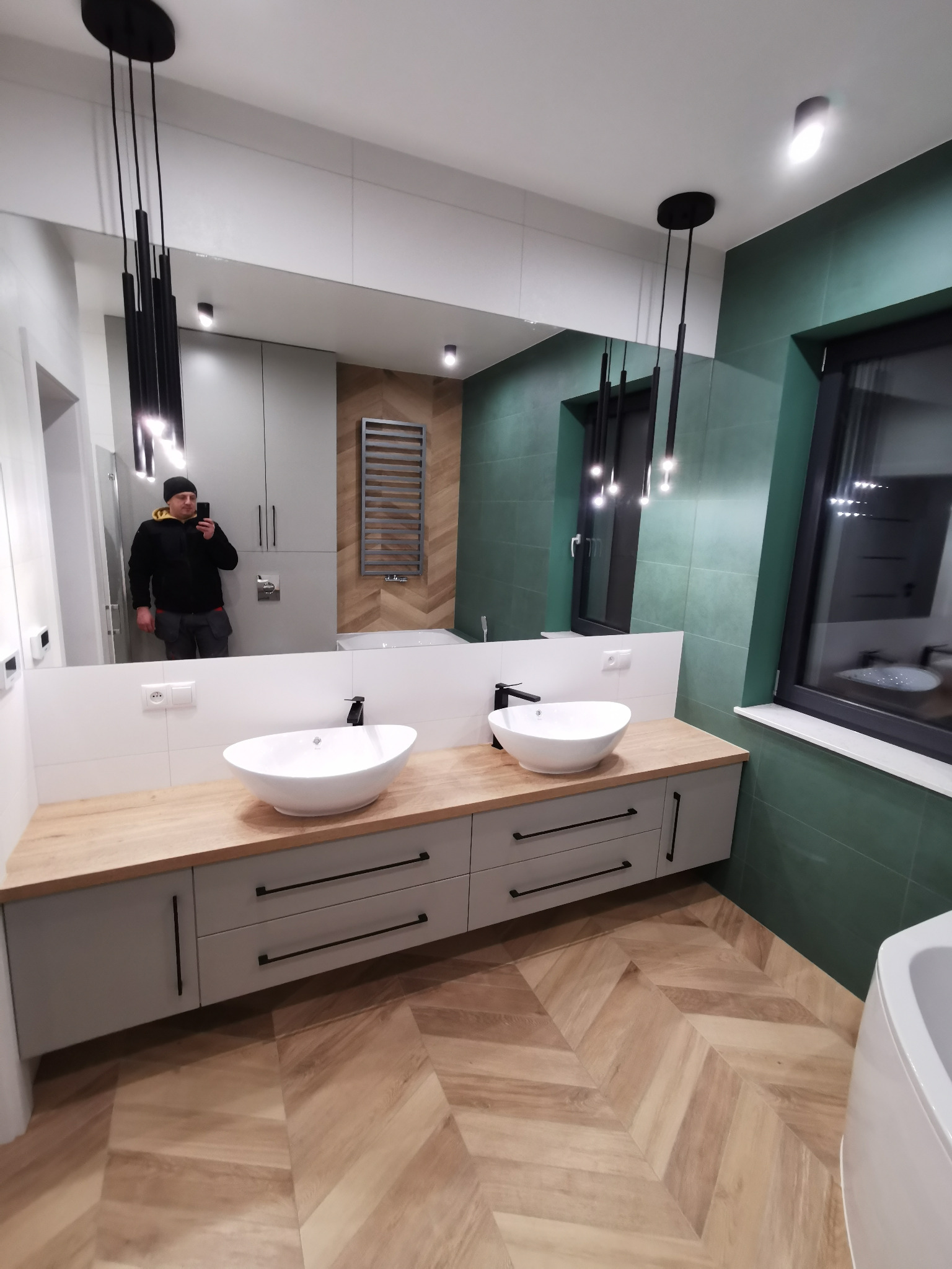 Nowoczesna łazienka z dwoma owalnymi umywalkami na drewnianym blacie, szare szafki z długimi uchwytami, zielone płytki na ścianach, podłoga ułożona w jodełkę, czarne wiszące lampy i odbicie...