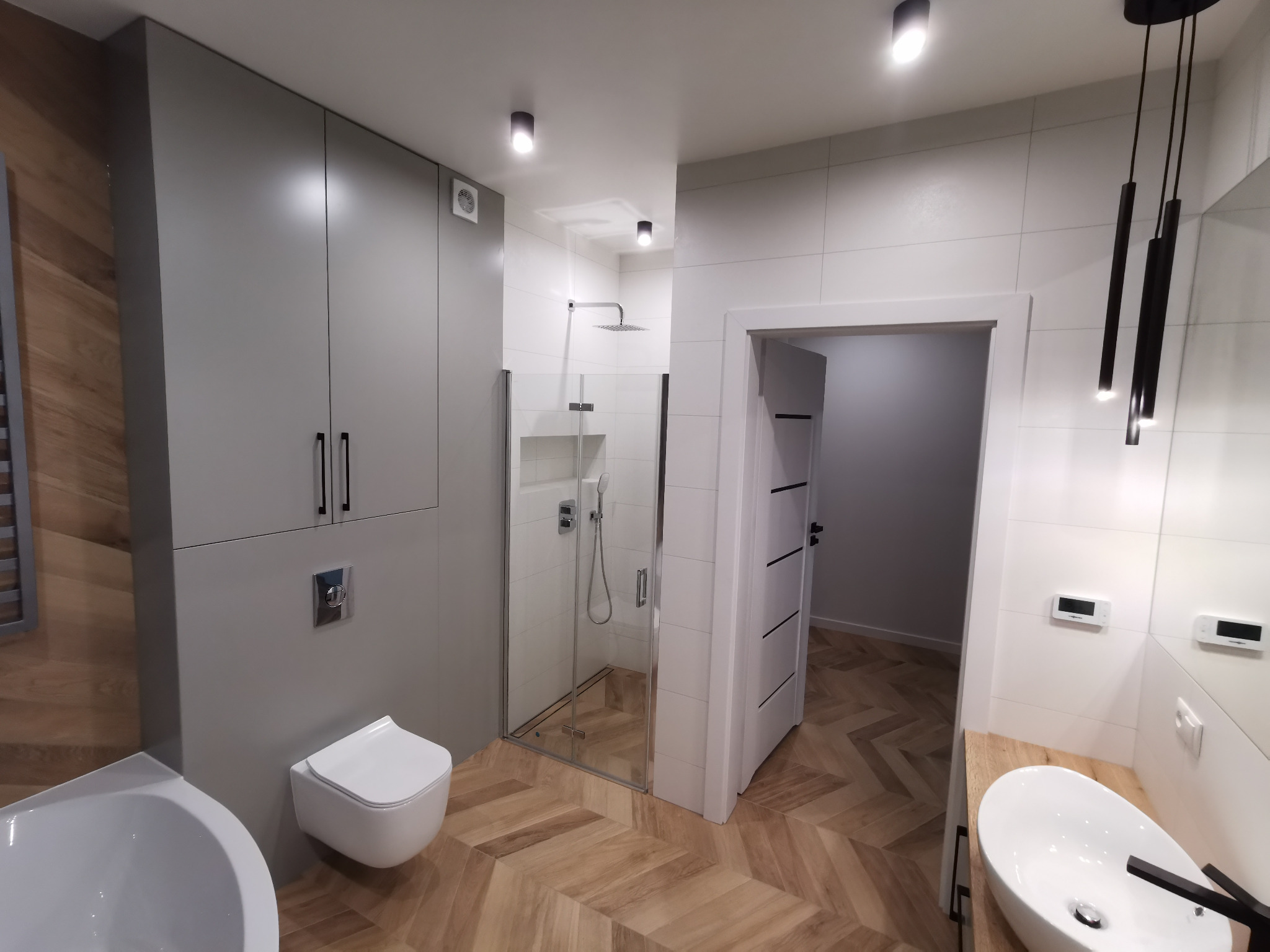 Nowoczesna łazienka z drewnopodobną podłogą w jodełkę, szarą zabudową meblową, kabiną prysznicową ze szklanymi drzwiami i wiszącą toaletą.
