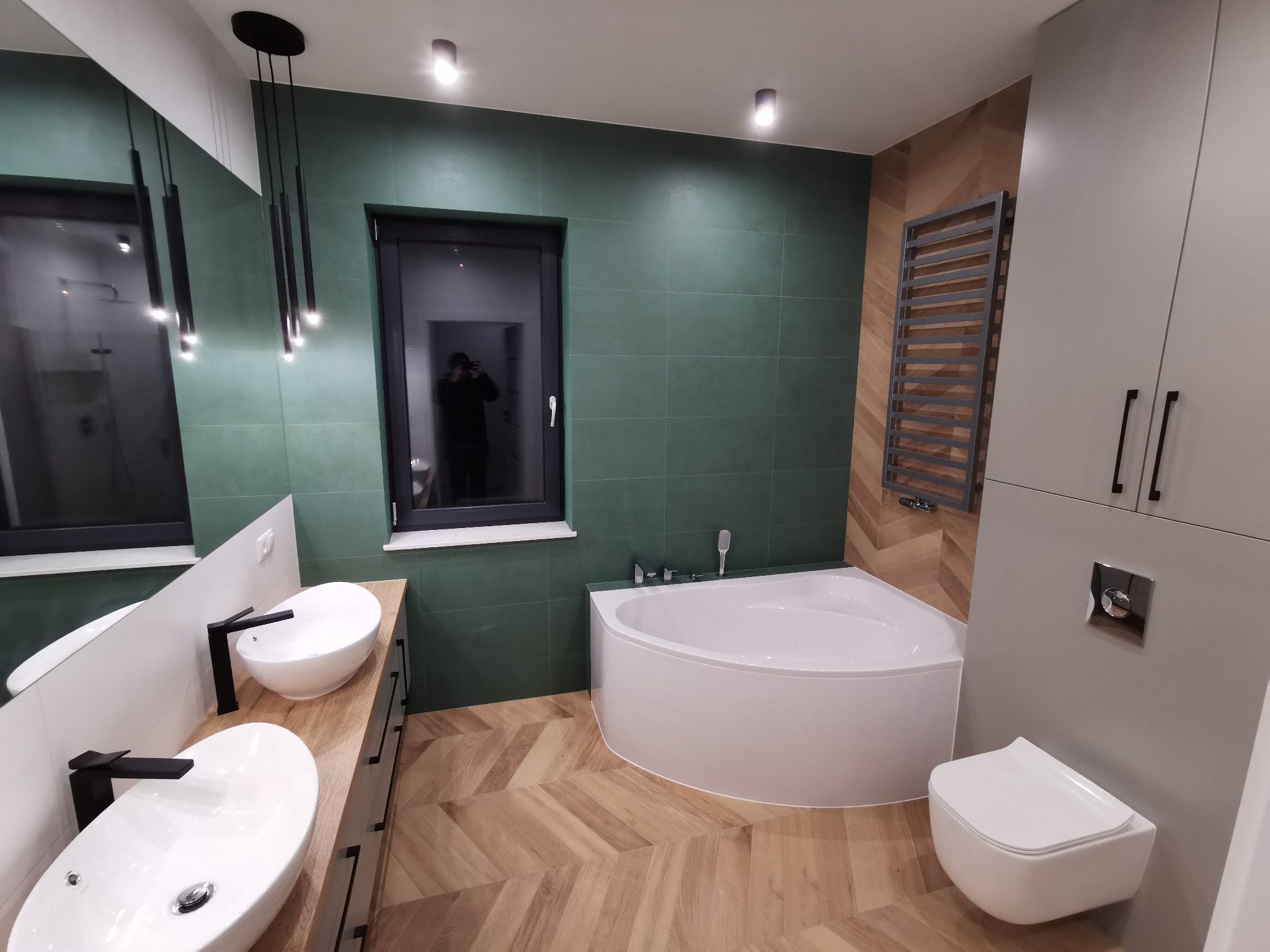 Nowoczesna łazienka z zielonymi płytkami, drewnianą podłogą ułożoną w jodełkę, białymi umywalkami nablatowymi i wanną narożną pod oknem.