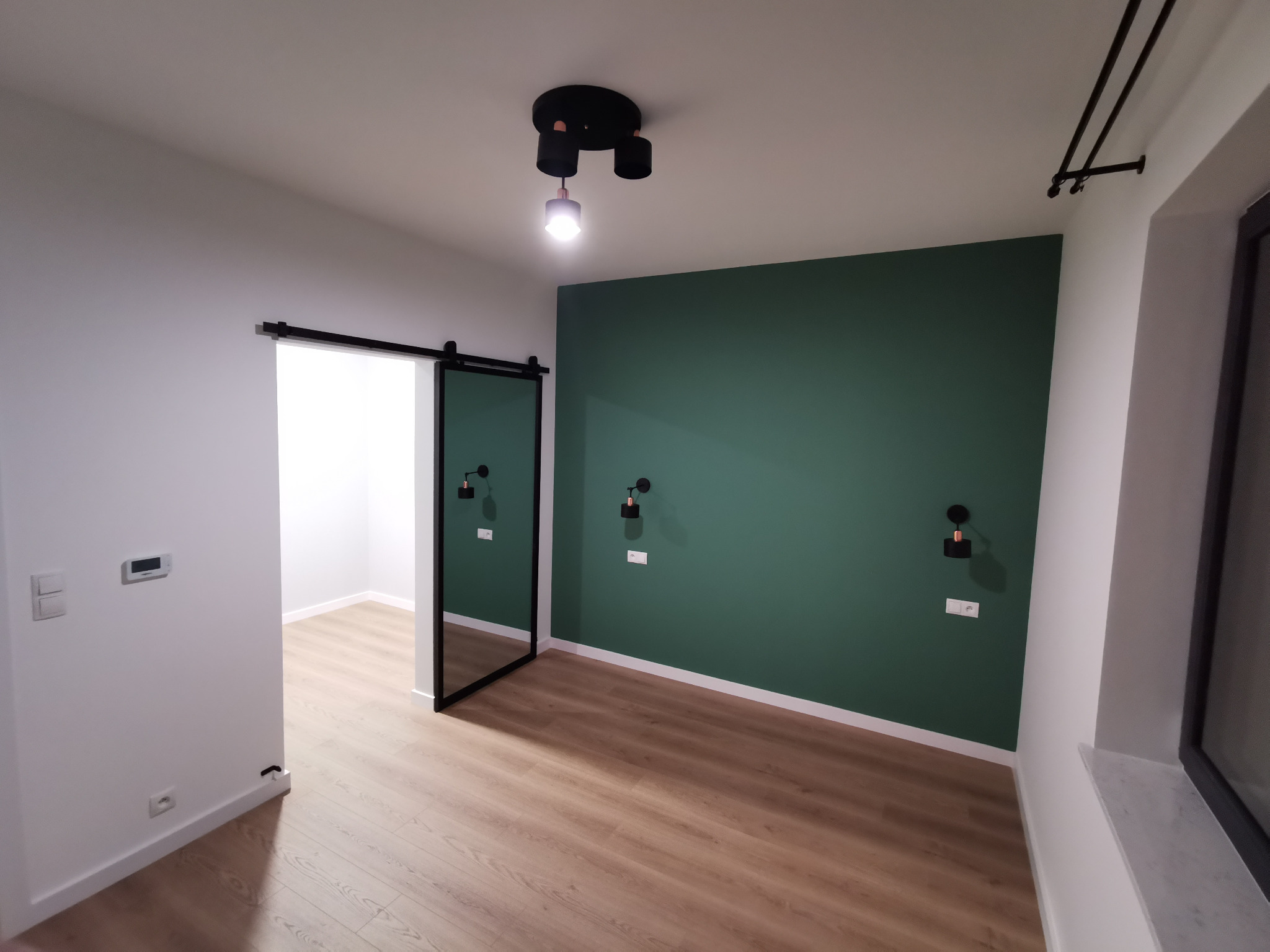 Nowoczesne wnętrze z zieloną ścianą, przesuwnymi drzwiami w czarnej ramie i drewnianą podłogą, oświetlone czarną lampą sufitową i kinkietami.