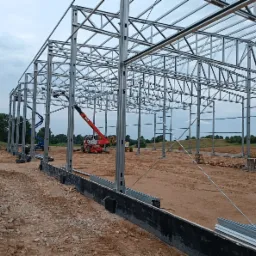 Montaż szkieletu stalowego hali na przygotowanym terenie, widoczny dźwig teleskopowy i podnośnik koszowy, w tle zieleń.