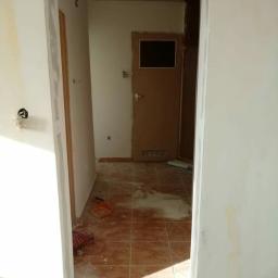 Marceli Iwanowski - Przygotowanie do malowania ościeżnicy drzwiowej w trakcie prac wykończeniowych, widoczne ślady szpachlowania na ścianie i brązowe płytki na podłodze.