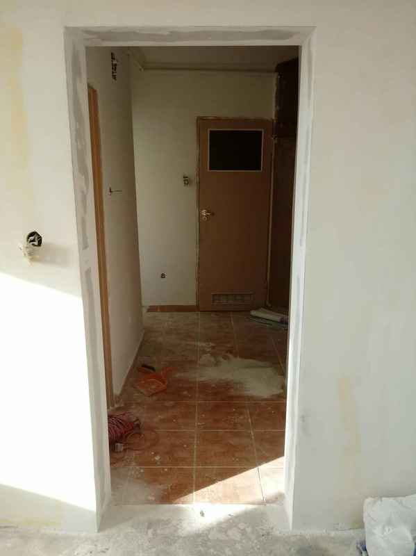 Przygotowanie do malowania ościeżnicy drzwiowej w trakcie prac wykończeniowych, widoczne ślady szpachlowania na ścianie i brązowe płytki na podłodze.