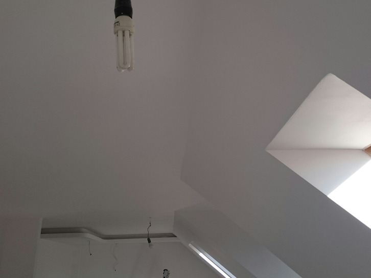 Biały sufit po malowaniu z widoczną lampą energooszczędną i oknem dachowym, fragment ściany i dekoracyjny element sufitu.