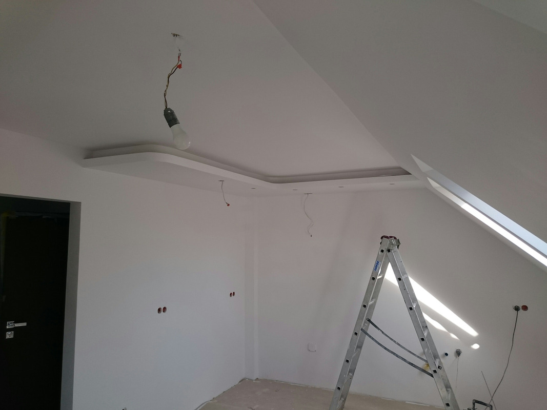 Pokój w trakcie malowania: białe ściany, sufit z dekoracyjnym obramowaniem, wisząca żarówka na kablu, aluminiowa drabina, widoczne puszki elektryczne.