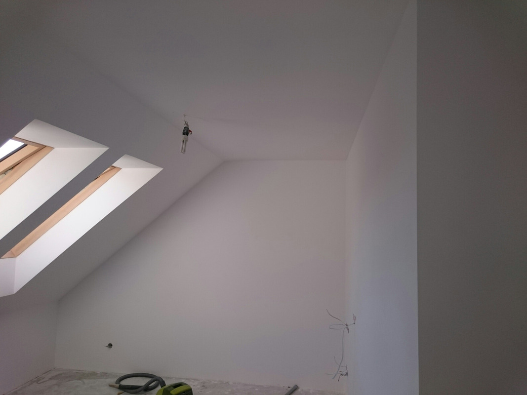 Poddasze po malowaniu na biało, widoczne okno dachowe z drewnianą ramą, wystające kable ze ściany i lampa wisząca na kablu, na podłodze leży odkurzacz przemysłowy.