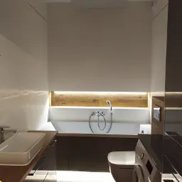 Nowoczesna łazienka z podświetlaną wanną, umywalką nablatową i zabudową meblową w ciemnych odcieniach, uzupełniona o pralkę i toaletę w tym samym stylu.