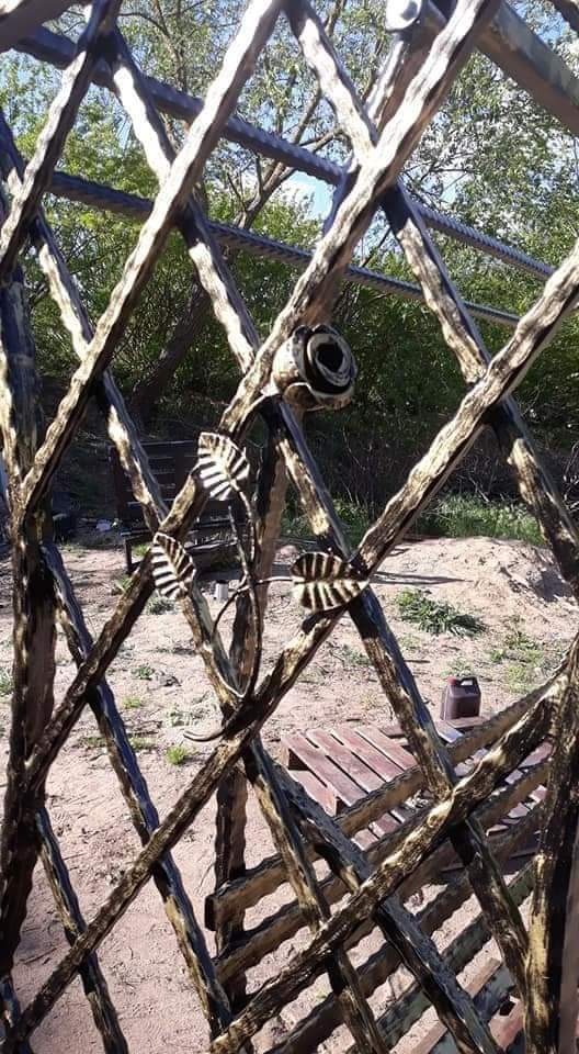 Dekoracyjne metalowe ogrodzenie z motywem kwiatowym i liśćmi, stylizowane na drewno, na tle ziemi i zieleni.